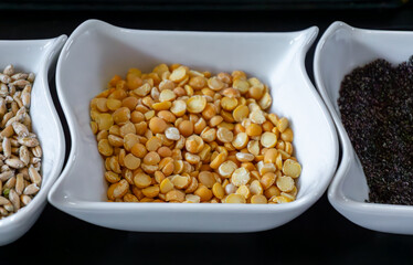 Close-up of yellow split peas in a white ceramic bowl placed on a black surface next to sunflower seeds and poppy seeds, kitchen ingredients