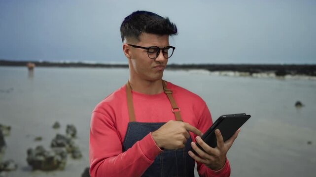 Man wearing apron holding tablet at seaside shows curiosity with thoughtful expression on beach with rocks in the background.