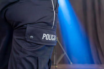 Navy blue uniform trousers of a Polish police officer with “Policja” label on side pocket, illuminated by blue light during a public service event © Adam