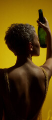 Back-view portrait of a pixie-cut woman in a shimmering backless gold dress, lifting a champagne bottle against a bold yellow studio background; glamorous celebration with dramatic rim light.