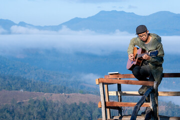 Asian male traveler relaxing and playing guitar outdoors during camping, enjoying peaceful nature and freedom in a beautiful natural setting.