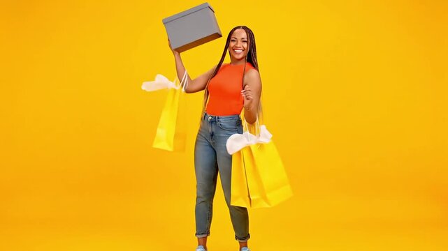 Joyful woman with a haul of shopping bags and a gift box, celebrating retail therapy success against a vibrant yellow background.