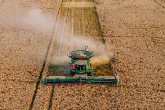 An aerial perspective shows a green combine harvester cutting through a golden wheat field, leaving parallel lines and a trail of dust in its path.
