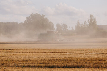 A combine harvester moves through a golden field, creating dust clouds. Distant trees and faint...