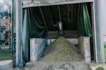 A storage facility with silage under a green tarp, concrete walls, and wooden beams. Industrial equipment and silos are visible to the left on dirt ground. © True Pixel Art