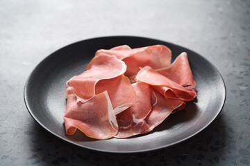 Thin italian prosciutto slices on black plate on conctere background