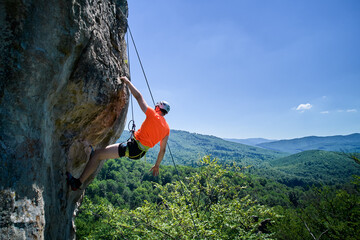 Aerial view of male rock climber ascending rugged limestone cliff with harness and rope for safety. Sportsman climbing on vertical large boulder at Dobvush Rocks in Carpathian mountains, Ukraine. © anatoliy_gleb