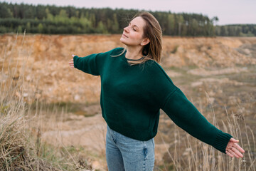 Joyful woman in green sweater with outstretched arms enjoying nature in open landscape, soft...