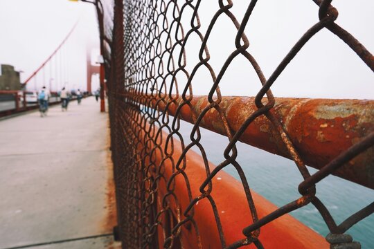 Corroded metal wire fencing on walkway toward Golden Gate Bridge, on a cloudy day, with fog on water