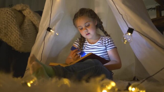 girl reading inside blanket tent with flashlight and book surrounded by fairy light and soft pillow child smiling cozy indoor bedtime reading in tent play with warm light and gentle expression