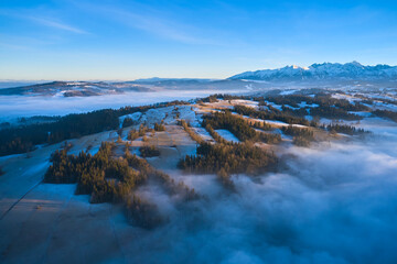 Fototapeta premium Drone view of Podhale highlands in Poland with winter frost, fog, and distant Tatra Mountains. Early morning light highlights the forested hills and valleys of this scenic mountain region.