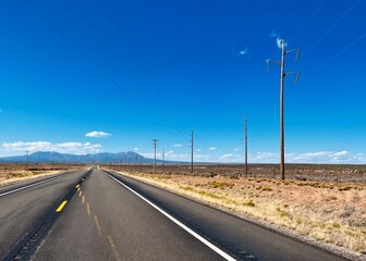 Fototapeta premium A straight desert highway in the American Southwest with uneven road markings, power lines, and distant mountains under a bright blue sky. Classic scenery of open road travel.