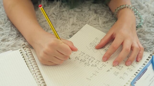 Close-up view of young student diligently solving complex math problems in spiral notebook, using pencil and calculator to check arithmetic homework and study for an exam