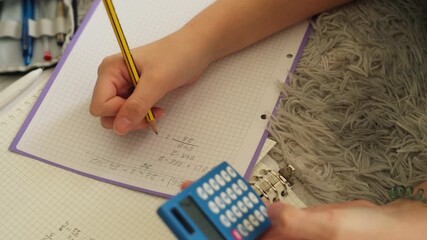 Child's hands writing down math equation on grid paper with pencil while simultaneously using small blue calculator to find solution, illustrating process of learning - Powered by Adobe