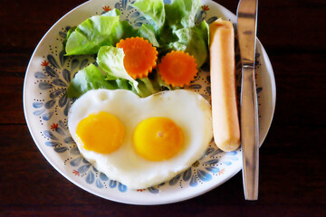 Heart shaped fried eggs with fresh vegetables and salad dressing and hot dog with knife in white dish 