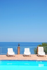 Minimalist summer scene with white deck chairs by a swimming pool overlooking the calm blue sea.