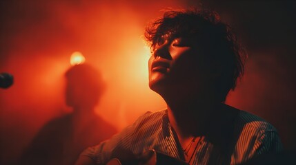 Young male singer with curly hair performs passionately on stage bathed in vibrant red light with musician.