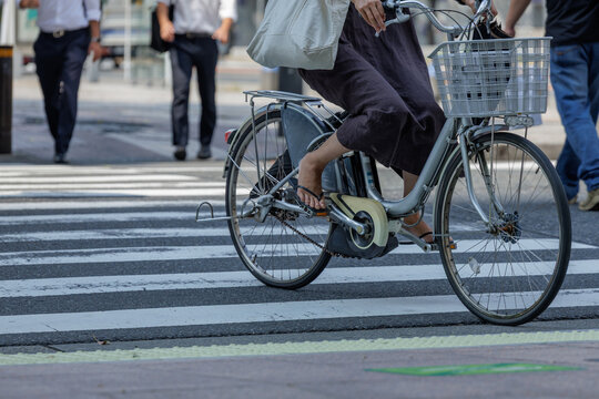 Cyclist rides across busy urban zebra crosswalk carrying bag and umbrella on bicycle with front basket, and passing by casually dressed pedestrian in jeans. Two business men visible in background.