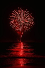 A vivid red firework bursts over a calm sea at night, casting a glowing reflection on the water. Silhouettes of boats dot the horizon, creating a festive coastal moment.