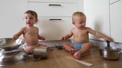 Baby and toddler play with pot pan and spoon on wooden floor in modern kitchen near cabinet while making playful mess with stainless bowl and utensil exploring texture and sound together joyful - Powered by Adobe