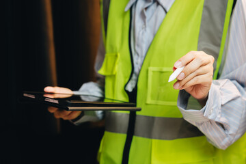 Professional Worker in Safety Vest Holding Tablet and Stylus in Industrial Setting with Focused Expression and Soft Lighting
