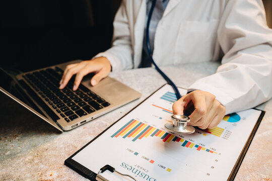 Doctor in white coat analyzing health data on laptop while holding stethoscope over medical report and charts