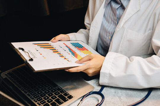 Doctor or scientist holding report with colorful graphs in a medical office setting, analyzing data on a laptop
