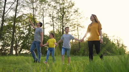 Family walking hand in hand through green grass at sunset in park, mother and father holding child and children, smiling and stepping across meadow, nature and tree line behind, relaxed family walk