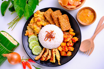 Top view of Indonesian Food, Nasi Uduk, with white rice, fried tempeh, spicy potatoes, and chili sauce, served on a black plate with fresh leaves and herbs surrounding it on a white background