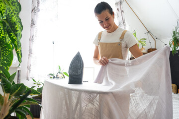 A housewife woman in an apron irons linen curtains in a room on an ironing board