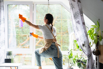 Woman in rubber gloves and apron manually washes window of house with rag cleaner and mop inside interior with home plants on windowsill. Restoring order and cleanliness in spring, cleaning servise