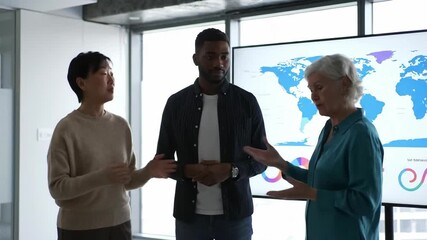 A diverse group of colleagues standing together inside a modern office space, looking toward a large display screen with world map data, discussing global strategies and future goals. - Powered by Adobe