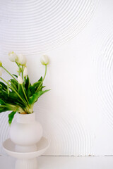 White vase with fresh tulips and green leaves placed on a minimalist pedestal against a textured white wall, creating a serene and elegant home decor atmosphere