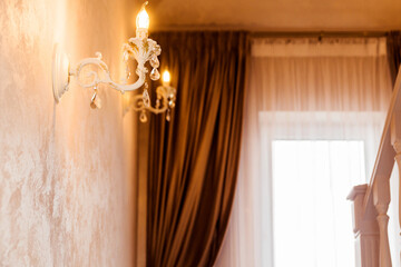 Elegant wall sconce with warm light illuminates a beautifully decorated interior space featuring rich drapery and soft textures, creating a cozy and inviting atmosphere