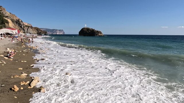 Sea beach waves crashing on shore with people and a cross on a rocky island under a clear sky on a sunny day