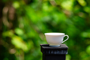 Single empty white ceramic coffee cup placed on top of a black iron fence post with blurred green foliage on the background.
