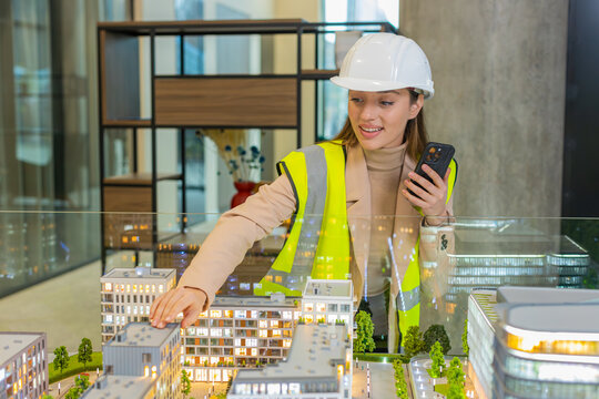 Confident young female architect in hardhat and reflective jacket using smartphone and analyzing business office complex architectural model. Woman girl smart designer of buildings working in office.