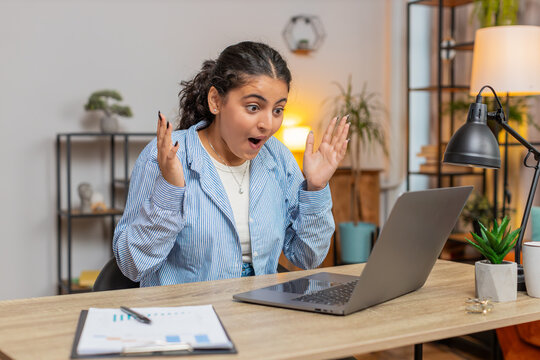 Happy young Indian woman working on laptop netbook shocked by sudden victory, winning, goal achievement, good career news. Excited freelancer girl clenching fists at home office workplace. Lifestyle.