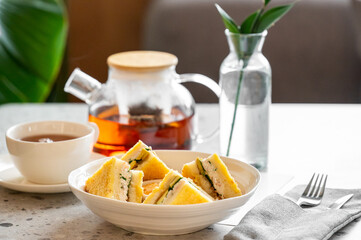 Breakfast table with triangular sandwiches, tea in cup and teapot, green plant in vase, and cutlery on napkin. Bright, clean, and cozy food presentation