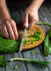 Hands are skillfully chopping green onions and leafy greens on a rustic wooden cutting board, with fresh ingredients scattered around in a cozy kitchen space