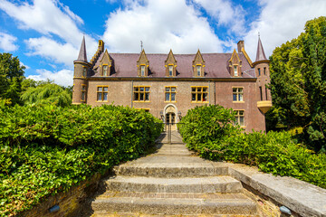 TerWormista Castle in frontal view, stairs leading to bridge over moat, brick walls, two side towers and gabled roof, surrounded by leafy trees, sunny day in Heerlen, South Limburg, the Netherlands