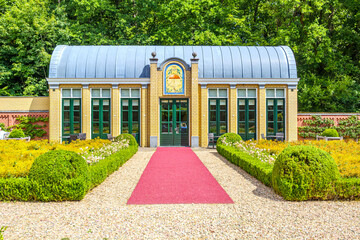 Front view of greenhouse in public park of TerWorm Castle, path with red carpet leading to entrance, leafy trees in background, yellow brick walls and gray roof, Heerlen, South Limburg, Netherlands