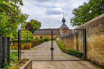 Modern metal gate with thin black bars, Cortenbach Castle with gabled roof and bulbous dome in background, green bushes and leafy trees, cloudy day in Voerendaal, South Limburg, Netherlands