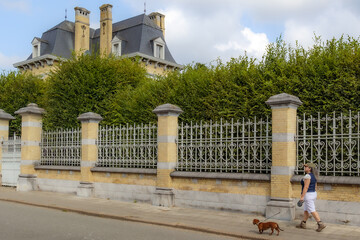 Senior woman walking with dachshund on sidewalk, brick wall with wrought iron outside the castle, leafy green trees, sloping hipped roof with chimneys in background in Rekem, Limburg, Belgium