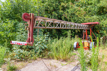Old, obsolete tractor with an agricultural crane, abandoned among lush wild vegetation of farm in agricultural land, rusty red metal arm, leafy trees in background, sunny spring day