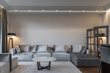 large modern living room, gray sofas and armchairs, gray curtains on large windows, coffee table, soft carpet, floor lamp in corner. room is flooded with bright light from window and from lamps.
