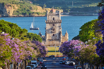 Scenic view of Belem Tower in Lisbon, Portugal, seen over a street with blooming purple jacaranda flower trees street with tourist sailboats on the Tagus River on sunset. Portugal
