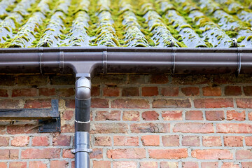 A brown plastic gutter and downspout on a rustic brick wall, below a mossy, corrugated roof. Highlights exterior building maintenance needs.