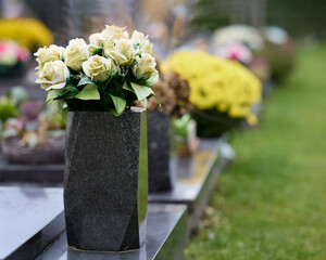 Elegant white roses in a dark stone vase adorn a headstone, set amidst other memorials and vibrant flowers in a serene, green cemetery landscape.