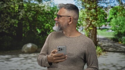 Man wearing glasses and sweater holding smartphone in forest, looking sideways and tapping screen with thumb near tree trunk; serenity reflection.